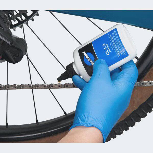 Park Tool CL-1.2 eco-friendly synthetic chain lube bottle, suitable for bike lubrication, with a workshop setting background.
